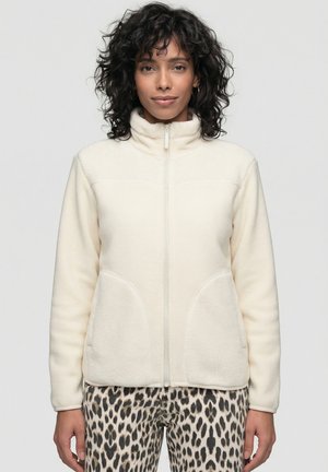 Frau mit lockigem Haar, die eine cremefarbene Fleecejacke trägt, die zugezippt ist, kombiniert mit Leoparden-Print-Hosen, stehend vor einem einfarbigen Hintergrund.