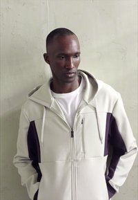 Sudadera con cremallera de color gris claro con un panel morado en la manga derecha, que cuenta con una capucha ajustable y un bolsillo delantero. Textura de tela suave.