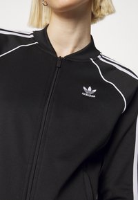 Svart Adidas zip-up jacka med vita ränder på axlarna och ärmarna, bärs av en person med kort blont hår och hoops-örhängen.