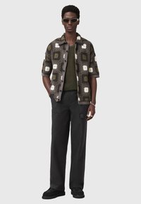 Camicetta a maniche corte, con un motivo a fiori in tonalità scure, abbinata a un top in maglia verde oliva e pantaloni scuri. Gli accessori includono occhiali da sole.