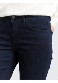 Vaqueros de mezclilla azul oscuro con corte slim, que presentan un bolsillo frontal y detalles en botón plateado, presentados sobre un fondo neutro.