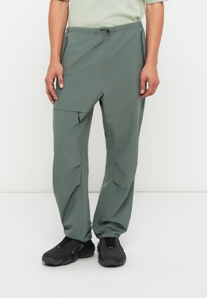 Πράσινο, ελαφρύ, κωνικό παντελόνι έχει ελαστική μέση, πλαϊνές τσέπες και λεία υφή, συνδυασμένο με μαύρες παντόφλες.