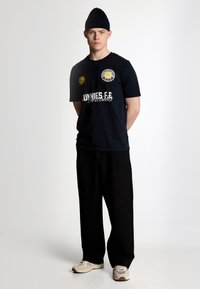 Marinblå t-shirt med kort ärm och gul emblem och text framtill, kombinerad med lösa svarta byxor och beige sportskor.