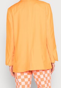 Oranger Blazer mit glatter Textur, strukturierten Schultern und einer einzigen Rückennaht, kombiniert mit karierten pastellrosa und orangen Hosen.