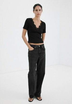 Woman wearing black lace-trimmed short sleeve top, high-waist wide-leg black jeans, and black flip-flops, standing with hands in pockets.