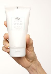 Un tube en plastique blanc de nettoyant pour le visage tenu dans une main. L'étiquette présente des accents verts et un texte noir. Le tube a un capuchon rond blanc. 150 ml.