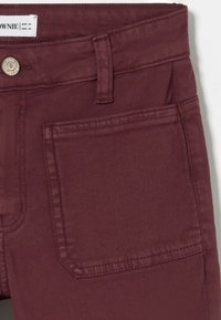 Primer plano de pantalones color burdeos con bolsillo delantero, trabilla para cinturón y botón de metal etiquetado como "BROWNIE", mostrando la etiqueta de tamaño EUR 34.