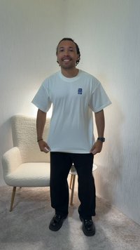 Άνδρας που στέκεται μέσα, φορώντας λευκό μπλουζάκι, μαύρο παντελόνι και μαύρα παπούτσια, με μια μπεζ πολυθρόνα και ένα μικρό στρογγυλό τραπέζι πίσω του.