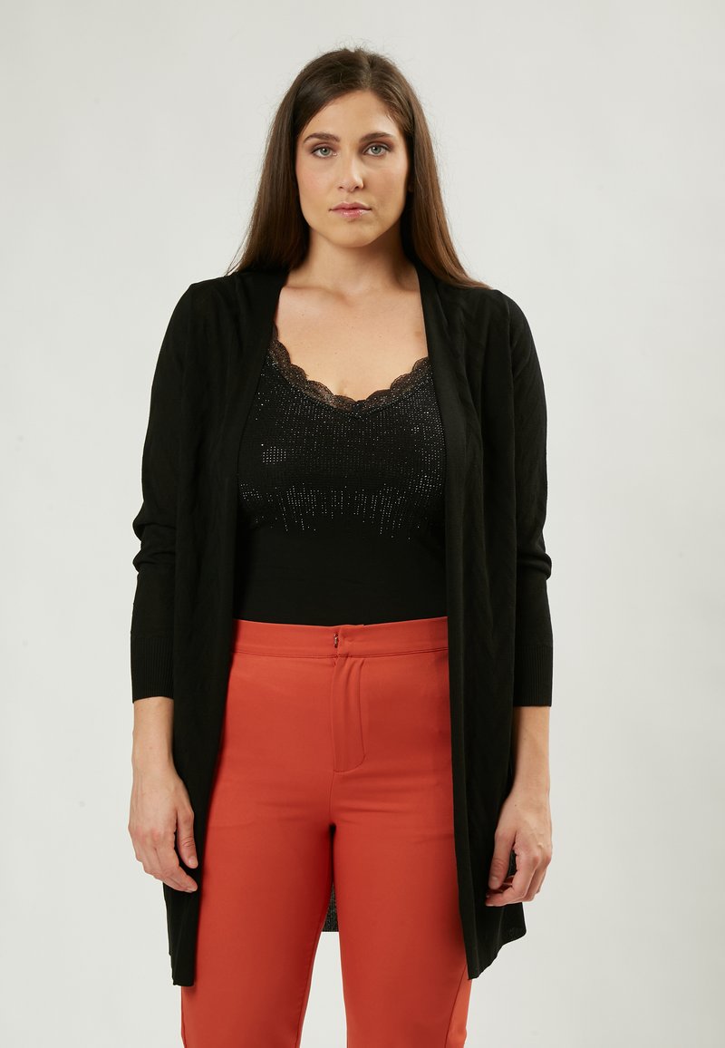 Schwarzer langer Cardigan mit strukturiertem Muster, offener Vorderseite, langen Ärmeln, über einem schwarzen Paillettenoberteil und orangen Hosen getragen.