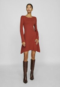 Robe à manches longues couleur rouille avec un col rond, un corsage ajusté, une jupe évasée et une texture lisse, accompagnée de bottes brunes montantes.