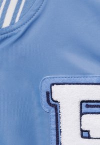 Gros plan sur un tissu bleu clair avec un écusson lettre en chenille blanche et bleu foncé, partie d'un vêtement avec un col rayé bleu et blanc.