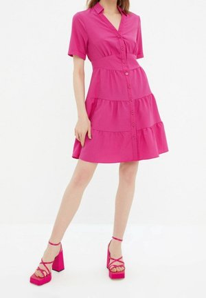 Kobieta ubrana w jasnoróżową, warstwową sukienkę na guziki z krótkimi rękawami oraz pasujące różowe platformy na obcasie z paskami wokół kostek.