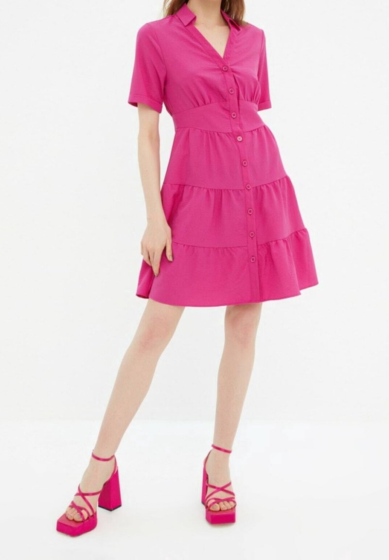 Femme portant une robe à volants boutonnée de couleur rose vif avec des manches courtes et des talons compensés rose assortis avec des brides à la cheville.