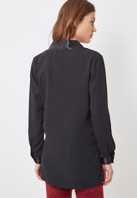 Camisa negra de manga larga con una textura suave y un cuello brillante. Presenta puños con botones y un dobladillo curvado. Modelo de espaldas.