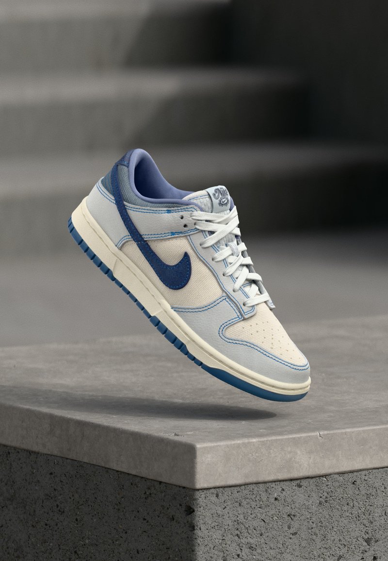 Zapatilla Nike blanca y azul con cordones flotando sobre un bordillo de hormigón y escaleras desenfocadas al fondo.