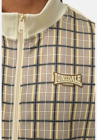 Jakke med lynlås i en beige, brun og gul geometrisk mønster. Har et tekstureret stof og et logo patches på venstre brystområde.