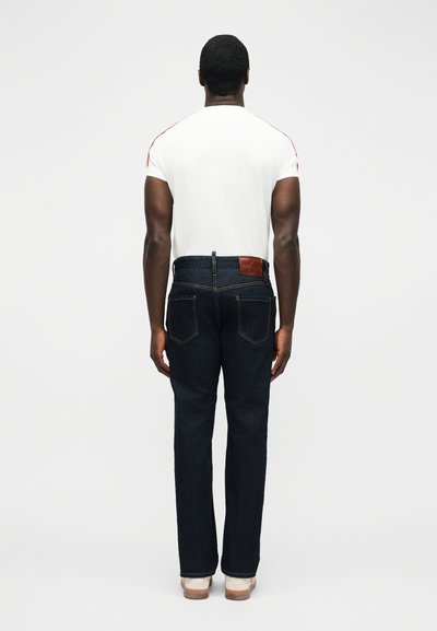 Άνδρας που στέκεται με την πλάτη γυρισμένη, φοράει λευκό κοντομάνικο πουκάμισο, σκούρα μπλε τζιν με πίσω τσέπες και λευκά παπούτσια με καφέ σόλες.