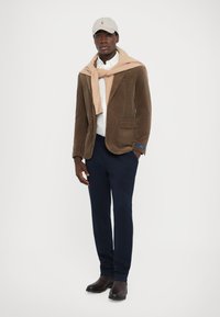 Blazer en velours côtelé marron, pull crème noué autour du cou, chemise blanche, pantalon marine, casquette grise et bottes marron foncé. Look propre et structuré.