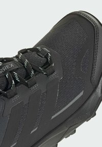 Scarpa da trekking nera con tomaia in mesh, punta rinforzata e suola in gomma testurizzata. Presenta lacci riflettenti e un distintivo design a tre strisce.