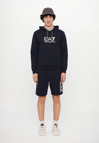 Navy blue hoodie with a kangaroo pocket and drawstrings, paired with matching shorts featuring a logo design on the side. Tan sneakers.