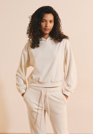 Donna con capelli ricci che indossa una felpa beige a coste con cappuccio e pantaloni coordinati, in piedi con le mani nelle tasche contro uno sfondo semplice.