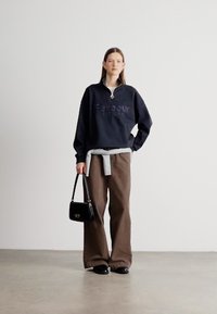 Μπλε ναυτικό φούτερ με γιακά μισού φερμουάρ και ανάγλυφο λογότυπο, συνδυασμένο με φαρδιά καφέ παντελόνια και μαύρη τσάντα. Ομαλό ύφασμα και άνετη γραμμή.