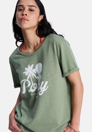 Frau mit dunklem Haar, die ein grünes T-Shirt mit weißem Palmenmuster und Text trägt, roter Bikini-Träger sichtbar, bunte Perlenarmband am Handgelenk.