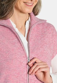 Gilet en tricot rose avec des côtes verticales, fermeture éclair et col haut. En dessous, une chemise blanche à col est visible.