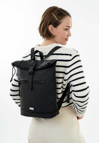 Sac à dos à rabat noir avec des bretelles ajustables, poche avant et fermeture éclair. Le matériau est lisse avec une finition mate. Design simple.