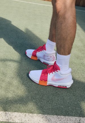 Baskets blanches de sport avec des accents roses et orange, en matière mesh, panneaux texturés, et un logo Nike visible. Portées avec des chaussettes blanches.