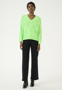Neon groene gebreide trui met een V-hals en geometrisch patroon, gecombineerd met hoge zwarte flared jeans en zwarte heeled enkellaarsjes.