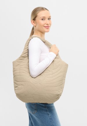 Woman wearing white long sleeve top and blue jeans holds large beige quilted tote bag over shoulder, smiling sideways.