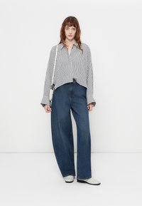 Gestreiftes, kurzes Shirt in Weiß und Schwarz; kombiniert mit weit geschnittenen, dunkelblauen Jeans; Modell trägt weiße Schuhe; schlichte Hintergrund.