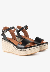 Sandalias de cuña de cuero negro con dos correas anchas, hebilla dorada y detalle de yute trenzado en la plataforma. Plantilla de cuero marrón.