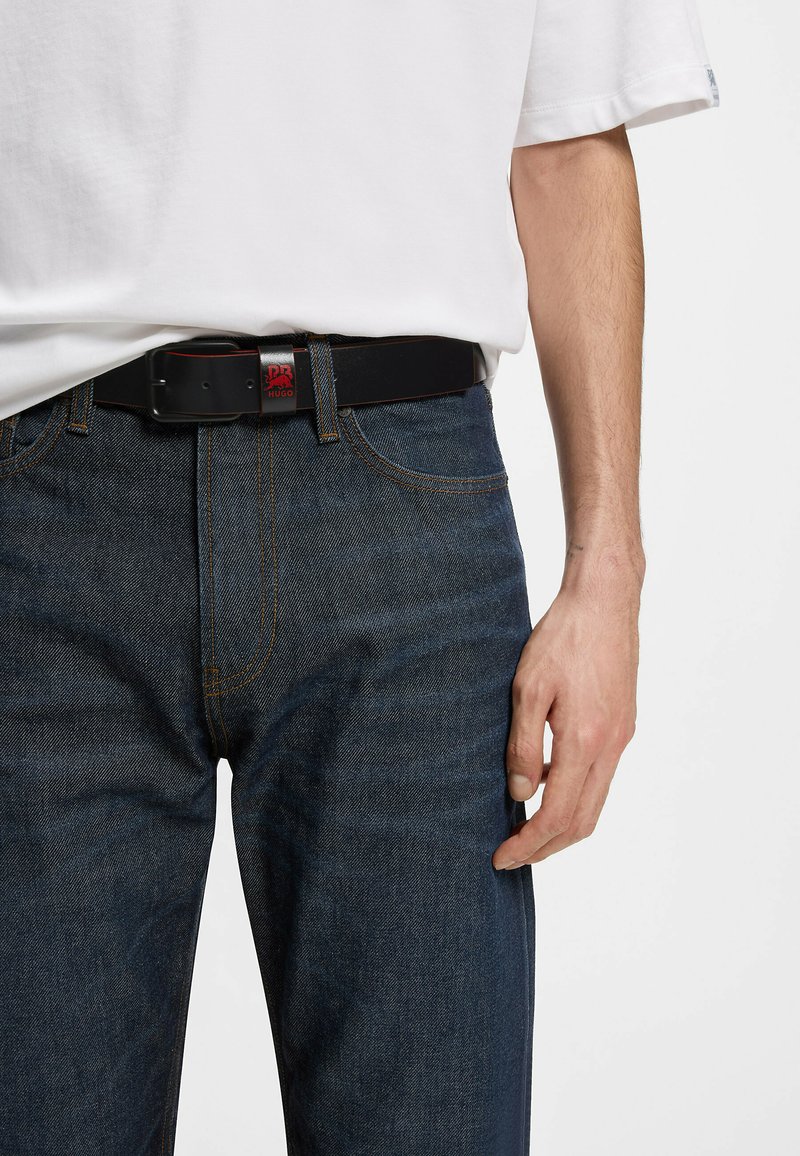 Adulte portant un jean bleu foncé avec des coutures orange, une ceinture en cuir noire avec un logo rouge, et une chemise blanche à manches courtes.