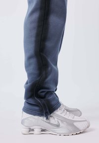 Persona que lleva pantalones de chándal azul oscuro con cremalleras en los tobillos y zapatillas Nike blancas con suelas de amortiguación visibles.