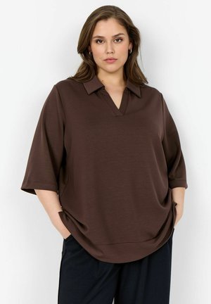 Femeie purtând o cămașă maro deschisă cu guler și mâneci până la cot, precum și pantaloni negri, stând cu mâinile în buzunare pe un fundal alb.