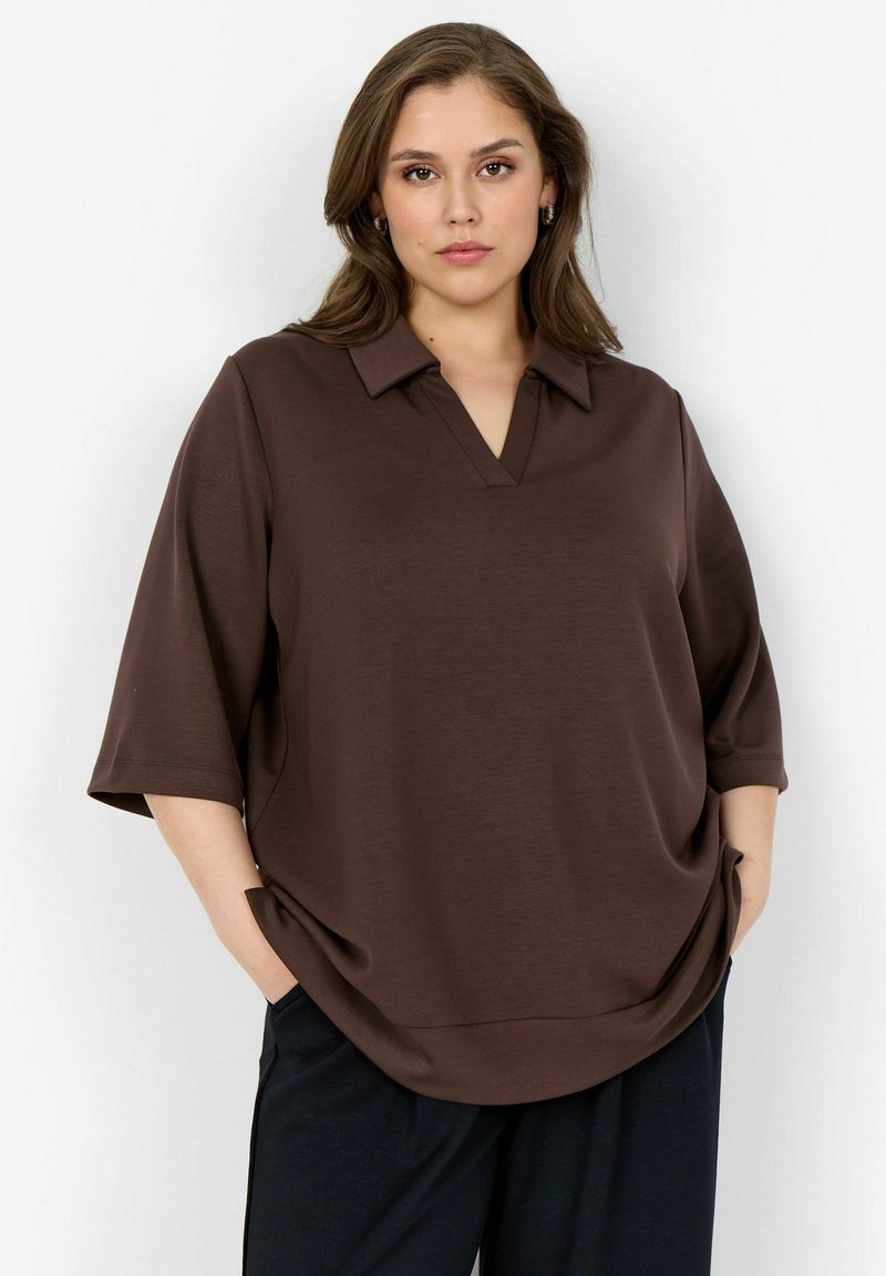 Frau trägt ein lockeres braunes Hemd mit Kragen und Ellbogen-langen Ärmeln sowie schwarze Hosen, steht mit Händen in den Taschen vor weißem Hintergrund.