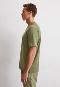 Olivegrünes T-Shirt mit kurzen Ärmeln aus weichem Stoff, mit Rundhalsausschnitt und lässigem Schnitt, seitlich an einem Modell präsentiert.