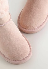 Pantofole in suede rosa con una consistenza liscia, punta tondeggiante e dettagli cuciti lungo le cuciture. Morbida rifinitura in pelliccia sintetica nella parte superiore.