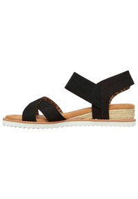Zwarte stoffen sandalen met kruislings gespen, tan kleurige binnenzool, espadrille-stijl hak met jute detail en een witte rubberen buitenzool. Verstelbare enkelriem.