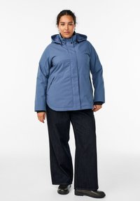 Šviesiai mėlyna vandeniui atspari striukė su kapišonu, priekiniais užtrauktukais ir kišenėmis su dangteliais, derinama su tamsiai mėlynomis plačiomis kelnėmis ir juodais batais.