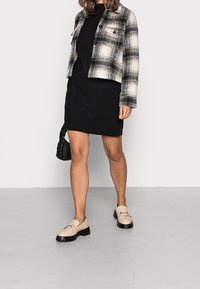 Rutigt yllejacka i svart och gräddfärg, parat med en figursydd svart klänning. Beiga loafers med svarta kraftiga sulor och en svart flätad handväska.