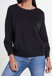 Schwarzer gerippter Pullover mit langen Ärmeln, Rundhalsausschnitt und geknöpften Manschetten, kombiniert mit blauen Jeans. Weiches Material mit lockerer Passform.