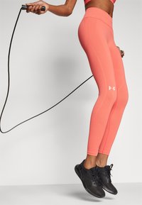 Korall színű leggingset és fekete sportcipőt viselő személy ugrókötelet ugrik egy sima világos háttér előtt.