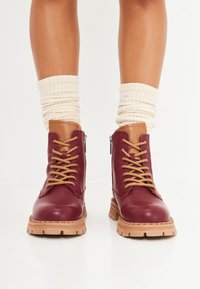 Botas de cuero burdeos hasta el tobillo con acentos en marrón claro, puntera redonda y cordones en contraste beige. Presentan una suela gruesa y texturizada y cierre de cremallera.