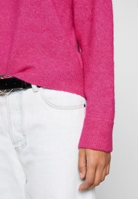 Suéter de punto fucsia con puños acanalados, usado sobre denim azul claro. El cinturón negro presenta una hebilla circular, creando un look casual.
