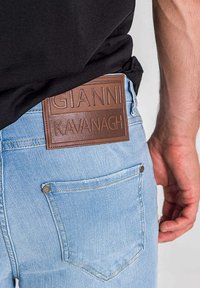 Jeans de mezclilla azul claro con una etiqueta de cuero marrón grabada con "GIANNI KAVANAGH" en el bolsillo trasero. Primer plano de la textura de la tela.