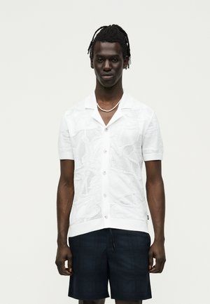 Homme aux cheveux tressés portant une chemise blanche à manches courtes avec des motifs texturés et un short foncé sur un fond uni.