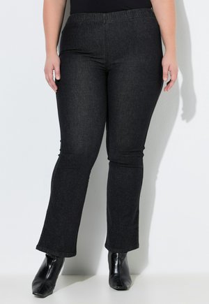 Kvinde iført sorte stretch bootcut jeans med elastisk linning og sorte ankelstøvler med hæl, stående mod en hvid baggrund.